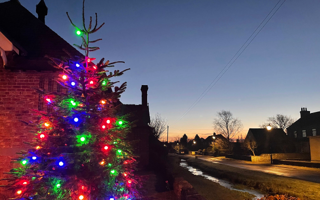 To the left of the picture is a Christmas tree with colourful lights. The sky is dark blue with the sun rising in the distance.